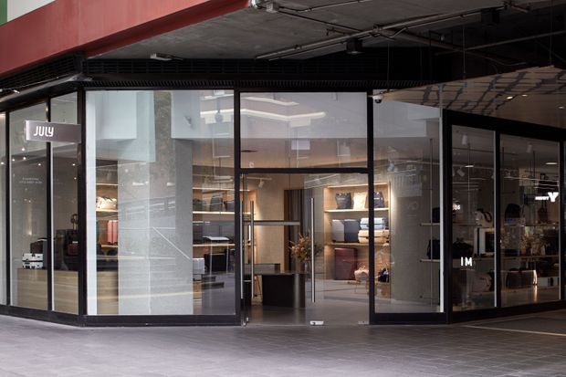 Street-level view of a contemporary retail storefront in a covered commercial arcade. The shop has extensive black-framed floor-to-ceiling glazing, glass entry doors, a dark exposed services ceiling, and a red fascia above. Warmly lit interior displays and shelving are visible through the glazing.