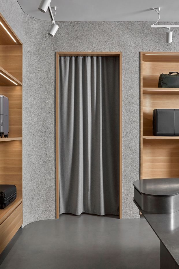 A compact, contemporary retail or showroom-like interior with a central curtained doorway, grey textured wall finish, timber-framed opening, built-in light timber display shelving, dark stone-look counter, and exposed white track spotlights. Suitcases or bags are displayed on the shelving, suggesting a product showroom or boutique retail space. The palette is predominantly grey, natural timber, and black.