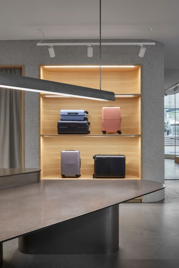 Contemporary retail interior featuring a sculptural dark reception or display counter in the foreground, a softly illuminated timber-lined shelving niche displaying suitcases, polished concrete flooring, grey textured walls, and track and linear pendant lighting. Glazing and additional retail space are visible to the right.