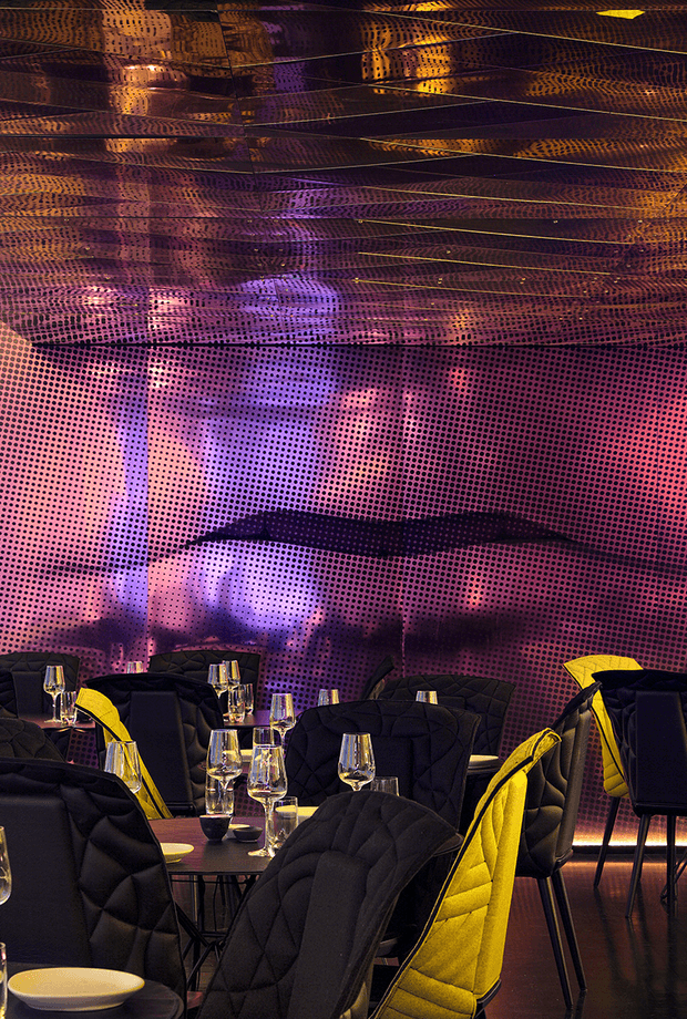 A dramatic, darkly lit contemporary dining or hospitality interior with rows of black quilted upholstered chairs, small round tables, and wine glasses. The walls and ceiling are covered in a reflective perforated patterned surface illuminated with vivid purple, magenta, amber, and blue lighting, creating an immersive, futuristic atmosphere. Bright yellow chair accents provide strong contrast.