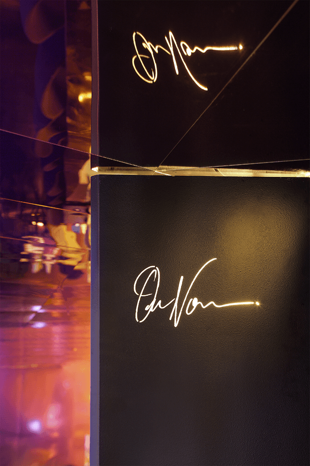 Close-up of a dark, textured feature wall with two glowing handwritten-style neon signature lights. Reflective panels at the left and along the upper section create purple, amber, and gold reflections, giving the installation a dramatic, high-end hospitality or entertainment ambience.