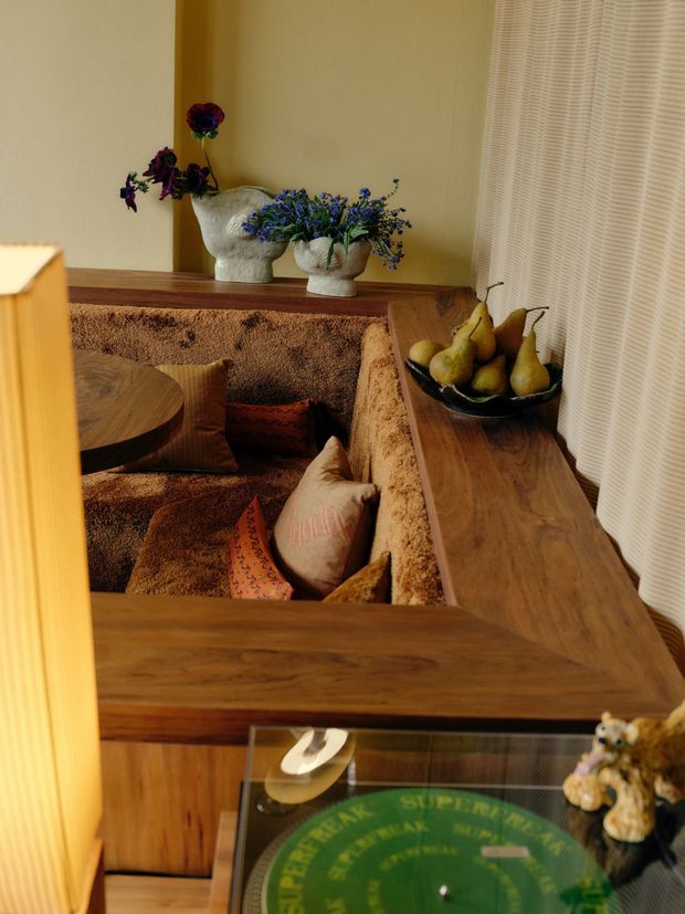 A warmly styled interior corner featuring a sunken conversation lounge enclosed by a thick textured brown upholstered bench, with patterned cushions and a round wooden table. A broad timber ledge and cabinetry frame the seating area, topped with sculptural ceramic vessels, blue flowers, and a bowl of pears. Cream pleated curtains and a mustard-toned wall create a rich, tactile, retro-inspired atmosphere. A glass-topped display table is visible in the foreground.