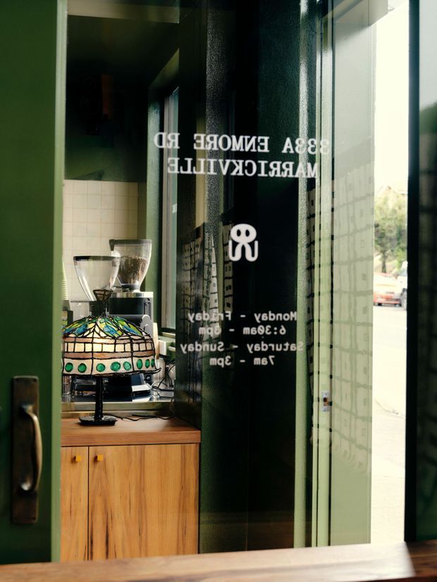Close-up view through a partially open glass entrance door into a small café or coffee shop. The foreground features dark green painted door and wall surfaces, a wood-framed glass door with reversed white opening-hours lettering, and a timber cabinet supporting a coffee grinder, espresso equipment, and a stained-glass table lamp. Bright exterior light is visible beyond the doorway.