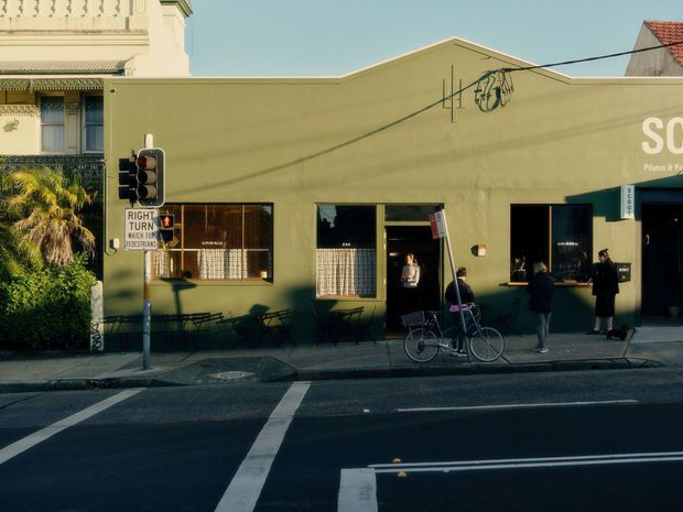 Street-level view of a low-rise commercial building with a muted olive-green rendered facade, simple pitched roof form, dark-framed windows and a restaurant or cafe frontage. Outdoor tables and chairs line the footpath, with pedestrians, bicycles, traffic markings and street signage in the foreground. A more ornate neighbouring building is partially visible at left.