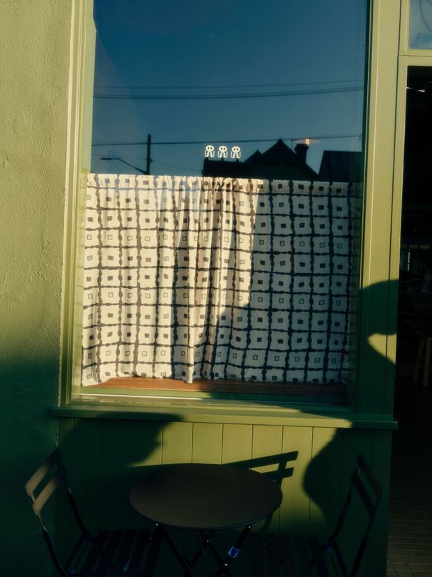 A close-up exterior view of a green-painted building facade with a large reflective window. The window has a patterned geometric curtain, with reflected rooftops and sky visible in the glass. A small outdoor cafe-style table and folding chairs sit below the window in deep shadow. The image has strong warm evening sunlight and high contrast.