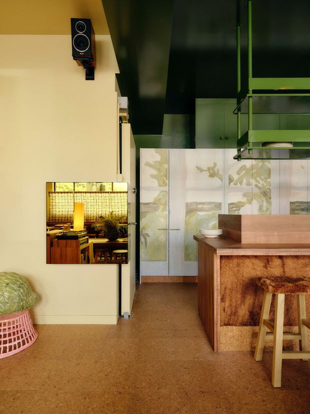 A richly styled contemporary kitchen and dining area with warm tan flooring, pale walls, dark green ceiling and cabinetry, and a timber island with a textured brown upholstered front. A large decorative tiled or painted panel forms the splashback, while a green-framed open shelf and wall-mounted television occupy the upper right. A reflective wall panel reveals a warmly lit dining setting with patterned wall treatment and timber furniture. The room has a playful, curated, retro-influenced atmosphere with strong material and colour contrast.