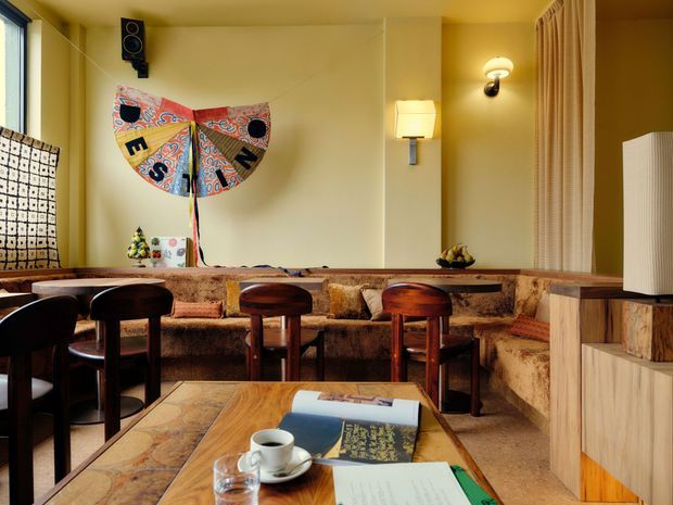 Warmly lit retro café or lounge interior with a long built-in upholstered banquette wrapping around the room, dark wooden stools and chairs, timber tables, a small service counter or cabinet at right, patterned curtains, wall sconces, and vintage artwork. The space has a richly layered, mid-century-inspired atmosphere with earthy mustard, brown, orange, and timber tones.