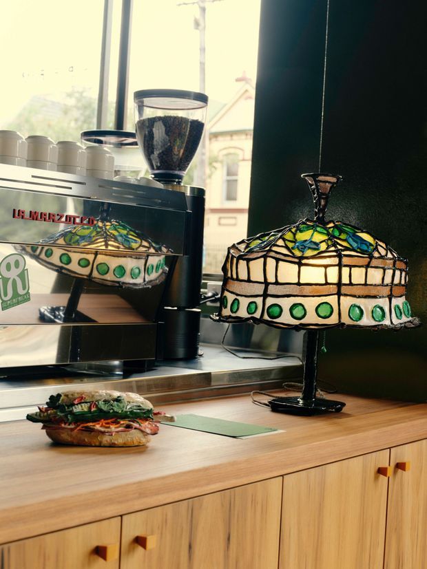Close-up interior view of a warmly lit café or kitchen counter featuring a black espresso machine with grinder, a colorful Tiffany-style stained-glass lamp, and a sandwich on a timber countertop. Light timber cabinetry with square wooden handles forms the lower portion of the scene, while a large window provides daylight in the background.