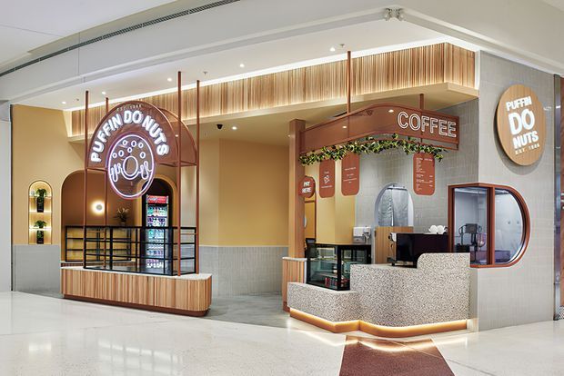 A contemporary indoor shopping-centre food and beverage tenancy featuring a Puffin Donuts counter and adjacent coffee kiosk. The fit-out includes curved timber-framed counters, terrazzo-look fronts, vertical timber slat detailing, warm integrated lighting, black metal screens, tiled lower walls, illuminated signage, and a polished light floor.