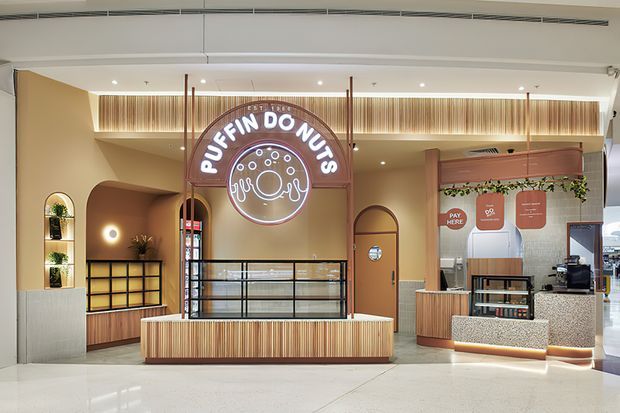 A contemporary commercial doughnut shop interior with a warm, refined fit-out. The storefront features curved architectural forms, ribbed timber-look wall and counter cladding, illuminated shelving, black metal-framed display glazing, integrated LED lighting, and a prominent circular neon “Puffin Donuts” sign. A service counter and coffee equipment are visible on the right, with pale terrazzo-like flooring and warm beige walls.