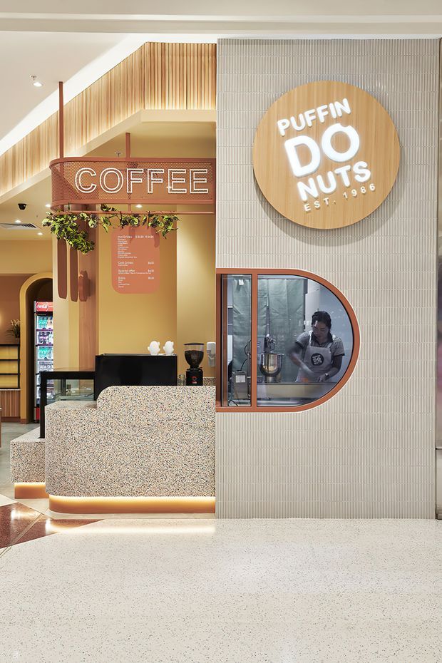 Interior view of a contemporary coffee and doughnut shop, featuring a terrazzo service counter with illuminated plinth, warm timber slatted architectural detailing, vertically ribbed light wall tiles, integrated menu signage, and a rounded service window into the preparation area. The fit-out uses warm wood, terracotta accents, cream surfaces, and playful branded lighting.