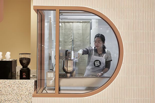 A contemporary café or commercial food-service interior viewed through a distinctive rounded glass service window. A barista works behind the counter with an espresso machine and mixing bowl, while the foreground includes a terrazzo counter, coffee grinder, and dark cabinetry. The service opening has warm timber-coloured framing and curved geometry, set against a vertically ribbed light wall finish.
