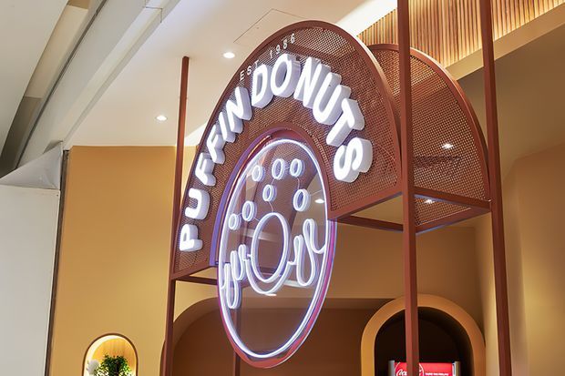 A close-up view of a commercial interior featuring a prominent illuminated “Puffin Donuts” sign mounted within a tall rust-red metal frame. The sign combines curved perforated metal panels, glowing white lettering, and a circular neon-style graphic. Warm peach walls, arched architectural openings, recessed ceiling lights, and vertical timber-look slats create a contemporary retail or food-service atmosphere.