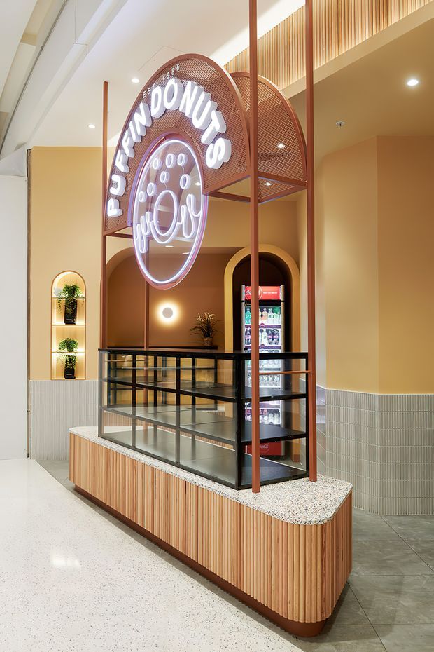 A warm, contemporary doughnut shop kiosk inside a commercial interior. The display features a curved terrazzo countertop, vertically slatted timber cladding, black metal and glass shelving, a refrigerated drinks cabinet, arched illuminated niches, and a prominent perforated metal canopy with neon doughnut signage. Pale peach walls, ribbed wall tiles, recessed lighting, and a polished terrazzo floor create a playful yet refined hospitality setting.