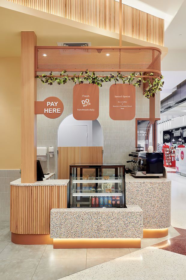 A contemporary commercial juice and snack counter in a shopping-centre setting. The compact kiosk features rounded terrazzo-clad counters with warm underlighting, vertical timber battens, pale green vertically tiled walls, a perforated curved copper-toned overhead canopy, hanging greenery, and prominent service signage. A refrigerated display case, coffee machine, point-of-sale equipment, and retail shelving are visible.