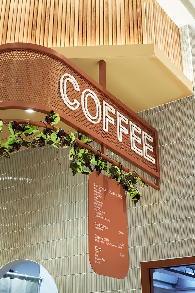 A close-up view of a café interior featuring a prominent curved terracotta-brown perforated sign reading “COFFEE,” suspended above a drinks menu. Vertical timber slats line the upper wall and balcony edge, while pale green vertically patterned wall tiles form the background. Hanging greenery, warm lighting, and rounded architectural forms create a playful, crafted hospitality fit-out.