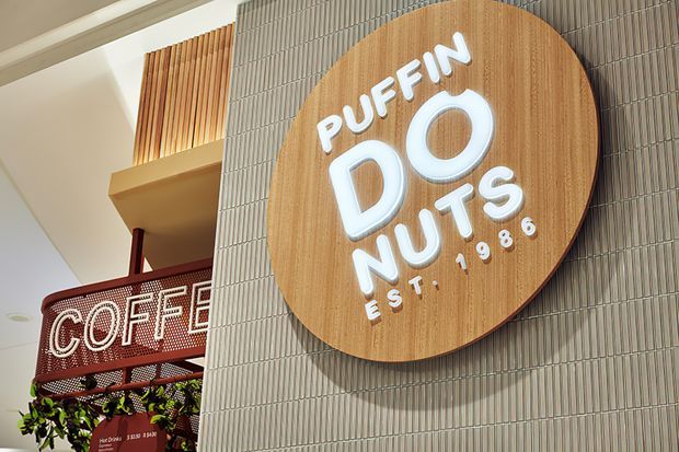 Close-up view of a commercial café interior featuring a prominent circular timber Puffin Doughnuts sign with raised white lettering mounted on a vertically ribbed grey tiled wall. A perforated red coffee counter and shelving with greenery are visible to the left, with warm timber slat detailing above.