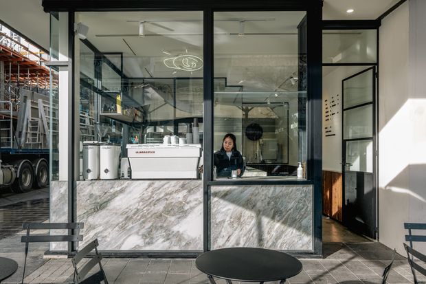 A compact cafe or commercial takeaway counter viewed from an outdoor seating area. The storefront has black metal-framed glazing and doors, a large polished veined stone-clad service counter, white walls, tiled flooring, and visible coffee equipment and shelving inside. Industrial warehouse shelving and a truck are visible through the glazing on the left, while outdoor tables and chairs occupy the foreground.