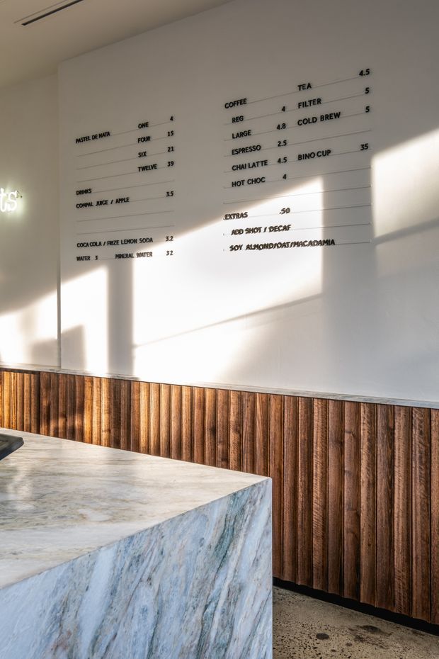 A sunlit cafe service area with a large pale veined stone counter in the foreground. The back wall features a white menu board with coffee, tea, drinks and extras listed in black lettering, while the lower wall is clad in vertically ribbed dark timber panels. The floor appears to be polished or sealed concrete, and strong window shadows create a warm, graphic atmosphere.