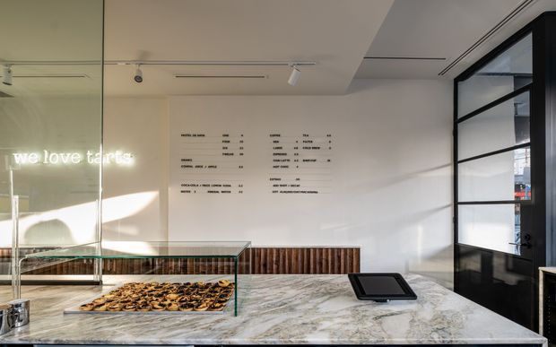 A bright, contemporary pastry or bakery counter viewed from the customer side. A large veined stone service counter displays pastries beneath a clear glass case, with dark wood vertical-panel joinery below and behind it. A white menu wall with black lettering, track lighting, and a black-framed glazed partition or door complete the refined commercial fit-out. A reflected illuminated sign reads “we love tarts.”