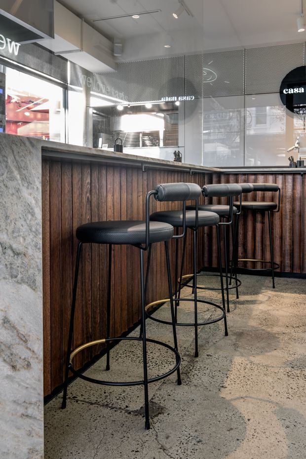 A contemporary commercial bar or café interior viewed from the customer side. The foreground features a long bar counter clad in dark vertical timber panels, topped with a grey stone surface, and lined with black upholstered counter stools with slim metal frames and brass-toned foot rails. The floor is a polished or sealed speckled concrete/terrazzo-like finish. Behind the counter are reflective glass and perforated metal screens, with integrated cabinetry and track lighting. The palette is dark timber, grey stone, black metal, and warm brass accents, creating a refined hospitality setting.