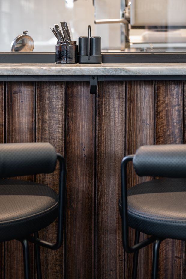 Close-up interior view of a compact kitchen or bar counter featuring vertically fluted dark timber cabinetry, a pale stone benchtop, matte black hardware and fixtures, and two dark upholstered bar stools. A reflective backsplash or mirror behind the counter shows the sink tap and countertop accessories.