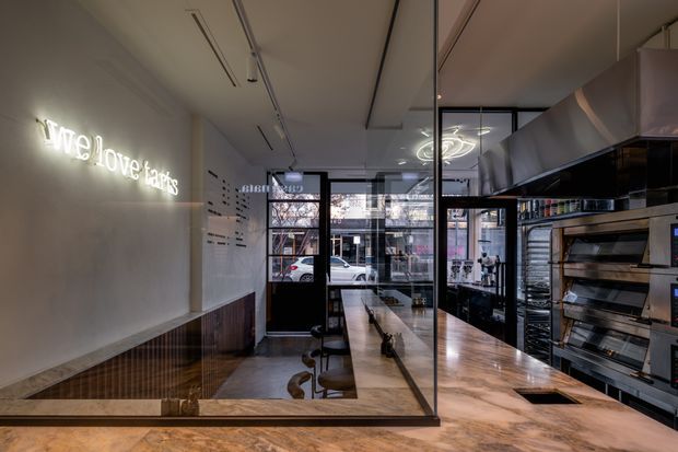 A narrow, contemporary commercial food-service interior, likely a bakery or café, viewed toward a glazed street frontage. A long timber counter with stools runs through the space, with commercial ovens, shelving, and stainless-steel equipment along the right wall. The interior features dark-framed glass partitions, warm timber finishes, a polished concrete or stone floor, linear ceiling lighting, and a glowing wall sign.