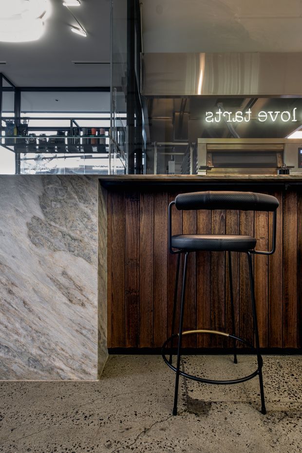 A close-up view of a contemporary café or bar interior, focused on a dark vertical timber-clad counter with a black upholstered bar stool. A large pale stone-clad counter or column occupies the left foreground. Behind the counter, black-framed glazing, exposed structural elements, shelving and reflected signage create an industrial hospitality setting. The floor is speckled concrete or terrazzo, with warm timber contrasted against dark metal and glass.