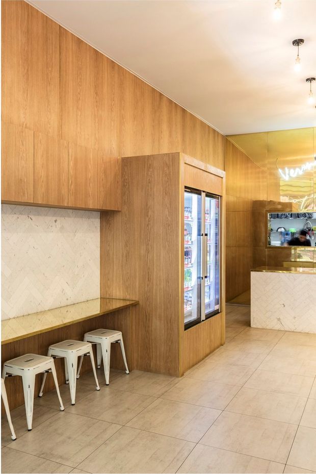 A warmly lit contemporary commercial café or dessert shop interior. The space features extensive vertical timber wall panelling and matching built-in cabinetry, pale marble-look tiled splashbacks and counter fronts, a tiled floor, a small built-in counter with three metal stools, refrigerated display units, and a service counter at the rear. Brass-toned wall panels and simple exposed-bulb pendant lights add a refined accent.