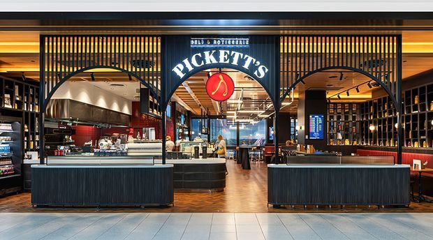 A polished commercial hospitality interior viewed from the front, with a prominent Pickett's sign above a wide entrance. The space features dark ribbed bar or counter fronts, black metal arched partitions with vertical slats, warm timber ceiling and flooring, illuminated shelving, display counters, bar seating, and warm ambient lighting. The open-plan venue appears to combine café, bar, and dining functions.