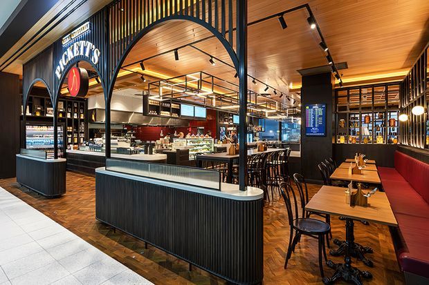 A stylish contemporary hospitality venue viewed from an open frontage, with a prominent coffee and food counter, illuminated display cases, high communal bar seating, booth seating, and extensive bottle shelving. The interior uses warm timber ceilings and parquet flooring, black metal arches and vertical screens, dark fluted counter fronts, pendant and track lighting, and deep red upholstered banquettes. The space appears to be a café, bar, or restaurant within a larger commercial setting.