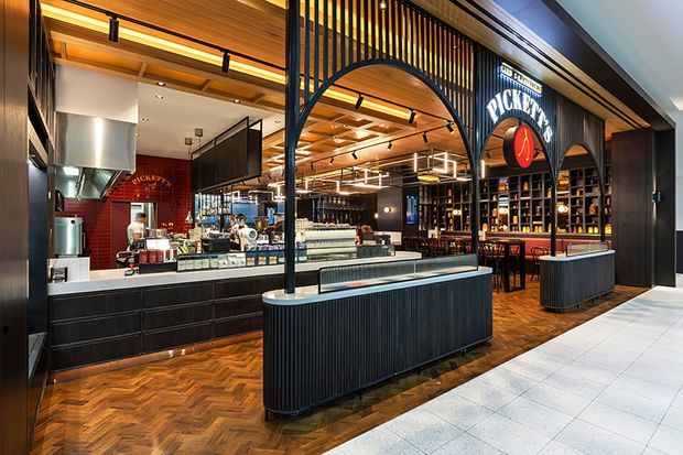 A sophisticated commercial hospitality interior, likely a coffee, bar, or restaurant venue. The open storefront reveals a service counter and commercial kitchen at left, a central refrigerated display counter, and a dark timber bar with extensive bottle shelving at right. Rich herringbone timber flooring, black ribbed joinery, arched black metal screens, warm timber ceiling lining, brass-toned details, and layered linear lighting create a refined, contemporary atmosphere.