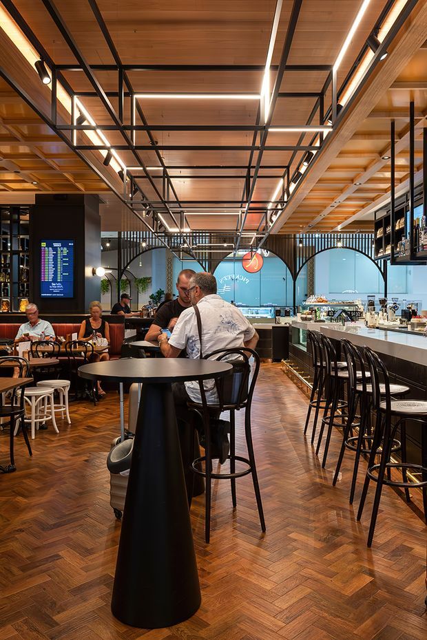 A warmly lit contemporary commercial dining and bar interior. The venue features a herringbone timber floor, timber-lined ceiling, exposed black metal ceiling grid and framing, linear LED lighting, arched glazed partitions, a long white bar counter, black bar stools, and dark round communal tables. Diners are seated throughout the space, with a digital display and shelving visible in the background.