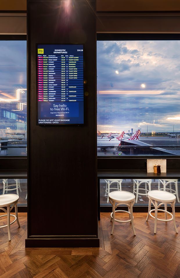 Interior view of an airport lounge or café overlooking an airport apron, with Virgin-branded aircraft visible through large windows. A central dark timber-clad column holds a flight departure display, while white bar stools line the glazing. The space has a warm herringbone timber floor, dark timber finishes, and contemporary airport hospitality furnishings.