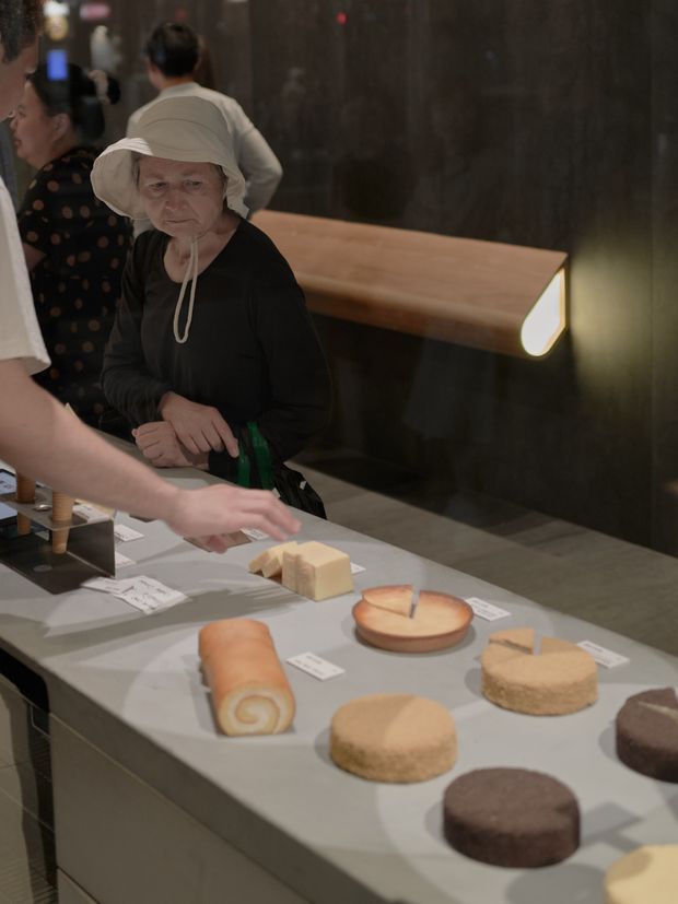A dimly lit commercial interior showing a food display or tasting counter with assorted cakes, pastries, and bread-like products arranged on a large light-coloured counter. Visitors stand around the counter, while a long suspended timber-toned feature light projects from a dark textured wall in the background.