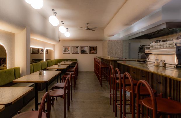 A warmly lit contemporary restaurant interior with a long open bar and kitchen counter on the right, upholstered banquette seating and marble-topped tables on the left, and rows of timber bar stools. Textured cream walls, framed artwork, globe pendant lights, dark polished flooring, and a ceiling fan create a refined, intimate hospitality setting.