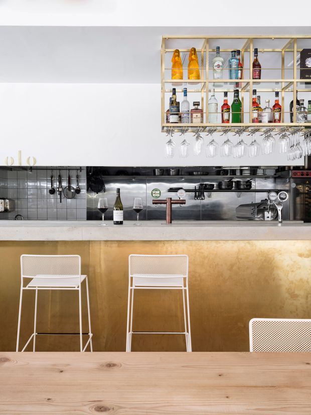 A stylish bar and hospitality counter viewed from the dining area. The scene features a brass-toned bar front, pale stone or concrete countertop, white mesh bar stools, exposed hanging glassware, open brass shelving stocked with liquor bottles, stainless-steel service equipment, tiled backsplash, and a timber dining table in the foreground. The overall design is clean, modern, and industrial-leaning with warm metallic accents.