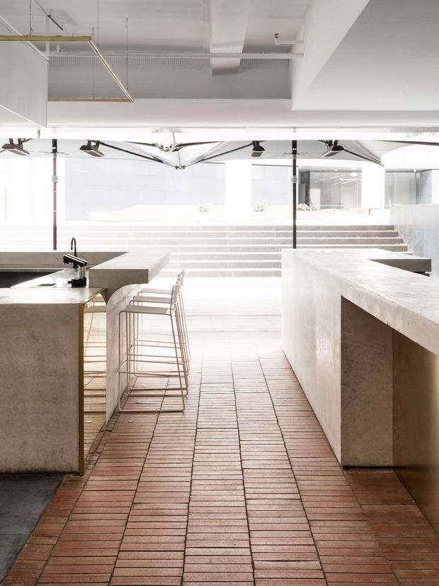 Bright, minimalist commercial hospitality interior with a central tiled walkway, large concrete service counters, a sink and tap, bar stools, and broad steps leading toward a glazed open area. The space has exposed ceiling services, suspended linear lighting, black structural poles, and abundant natural light.