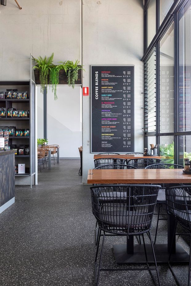 Interior view of a contemporary coffee shop or café with a tall industrial-style space, polished dark concrete floor, exposed concrete walls, full-height black-framed glazing, a large coffee menu board, built-in retail shelving, indoor plants, and timber communal tables with black wire-frame chairs.
