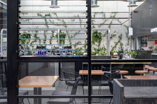 A contemporary commercial café or hospitality interior viewed through black-framed glazing. The space features a high glazed ceiling, exposed industrial-style pendant lights, dark tiled or stone-look walls, timber tables, black wire chairs, stools, a long counter, and abundant greenery arranged against a light gridded wall. The composition emphasizes the dining area and open, bright atrium-like setting.