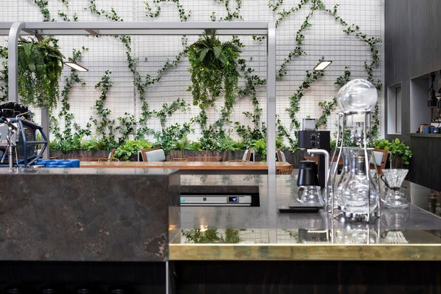 A sophisticated contemporary kitchen or hospitality-style bar area with a large dark stone island, integrated stainless-steel appliance, coffee equipment, and a reflective metal-topped counter. A white tiled wall with climbing greenery forms a striking feature backdrop, while a dark built-in joinery wall frames the right side. The space has a refined industrial character with strong material contrast, metallic finishes, and abundant planted greenery.