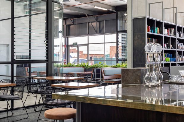 A bright industrial-style café or hospitality interior with a large polished stone or concrete-topped bar in the foreground, open shelving displaying glassware and crockery, and multiple timber tables with black wire-frame chairs. Full-height black-framed glazing and louvred windows bring in natural light, while exposed ceiling services, structural elements, and dark metal finishes reinforce the commercial warehouse character.
