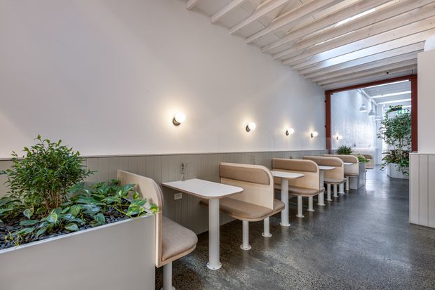 Bright, narrow commercial dining space with a row of built-in booth seating and small white tables along a paneled wall. The room features exposed white-painted ceiling joists, polished grey flooring, warm wall sconces, integrated planter boxes, and a glazed opening leading to a light-filled rear area with greenery.
