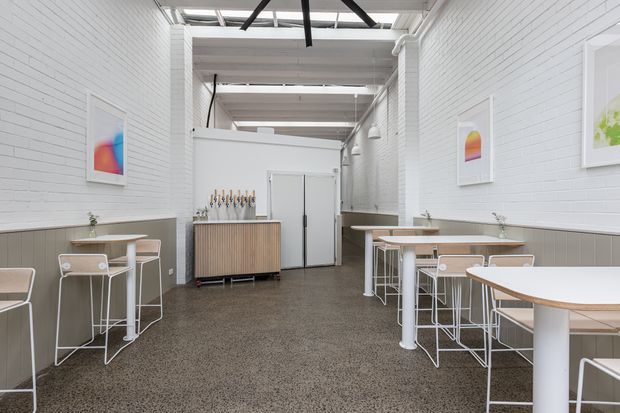 Bright contemporary commercial café or hospitality interior with white painted brick walls, a high exposed-beam ceiling and skylights, polished concrete flooring, multiple white high tables with stools, framed colourful artwork, and a timber-fronted service or storage cabinet. The space has an open, airy, minimalist industrial character.