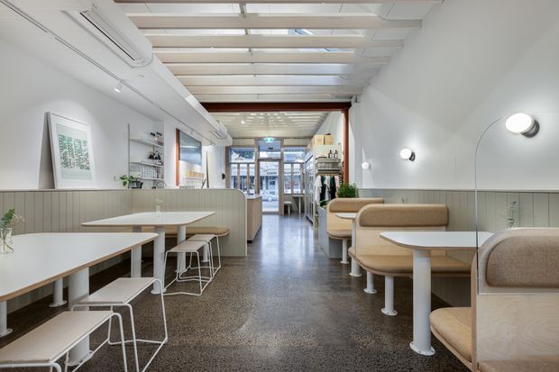 Bright contemporary café or hospitality interior with a long, narrow layout, polished concrete or terrazzo-style flooring, pale timber and white tables, built-in upholstered booth seating, a low fluted partition, wall shelving, and a glazed entrance at the far end. The ceiling features exposed white-painted beams and translucent roof panels, with simple wall sconces and linear air-conditioning. The space feels clean, airy, and inviting.