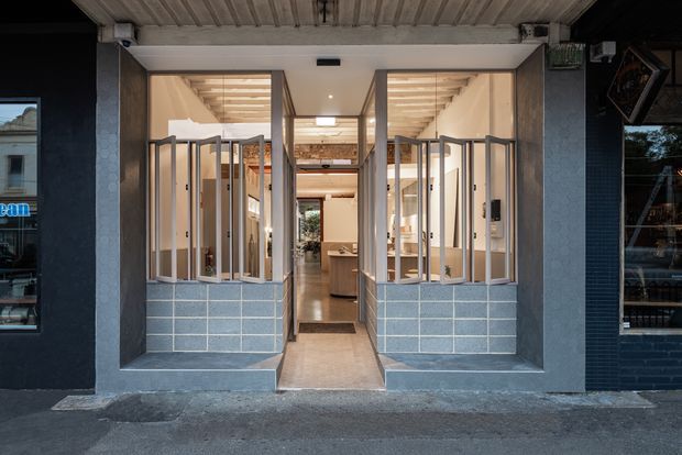 Street-facing view of a renovated commercial-style frontage with a central open entry and symmetrical glazed sections. The lower facade is clad in pale blue rectangular tiles, while dark painted masonry frames the opening. Inside, a warm timber-lined ceiling, exposed structural elements, and built-in joinery are visible.