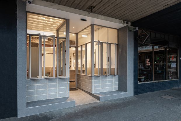 Street-level view of a contemporary commercial shopfront with a recessed glazed entrance and large steel-framed windows. The facade combines dark painted surfaces, exposed concrete framing, grey tiled lower walls, and a ribbed concrete soffit. Warmly lit interior shelving and display areas are visible through the glazing.