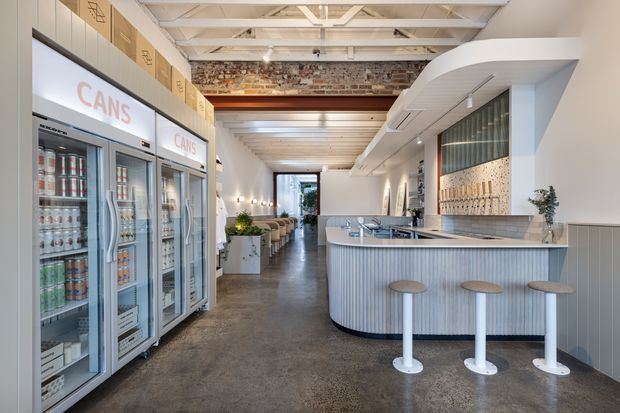 Bright contemporary commercial hospitality interior, likely a café or casual bar, with a curved fluted service counter, integrated sink and tapware, stainless-steel refrigerated display cabinets, bar stools, tiled wall detailing, exposed white-painted ceiling structure, and an exposed brick feature band. A seating area extends toward the rear glazing.