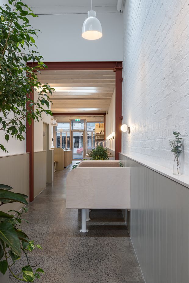 A bright, narrow commercial interior with a reception or workspace zone. The space features polished concrete flooring, white-painted brick walls, pale timber ceiling beams, exposed dark red structural framing, ribbed light-grey built-in cabinetry or counters, pale plywood workstation partitions, pendant and wall lighting, and indoor plants. Glazing at the far end opens toward an urban street.