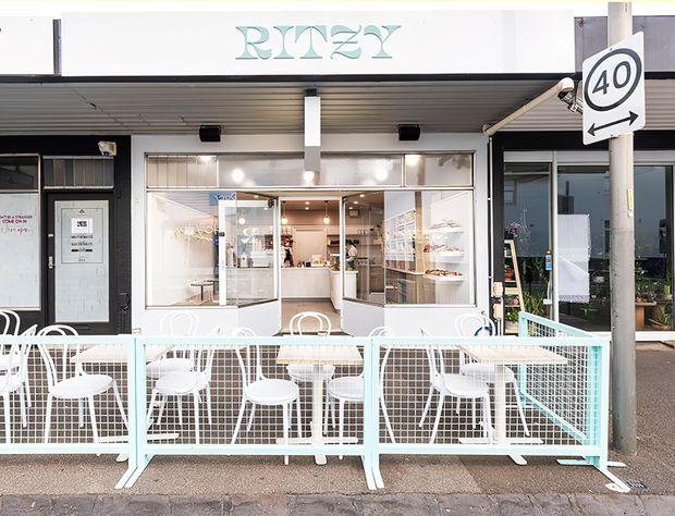 Street-level exterior view of a small contemporary café or food-service shop with a white and pale aqua storefront, glazed service frontage, covered awning, outdoor dining tables and white chairs enclosed by a matching mesh barrier. The sign reads “RINZY,” and a roadside 40 km/h sign is visible. The image primarily documents the commercial façade and outdoor seating area.